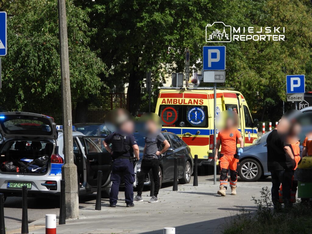 Ratownicy medyczni w pomarańczowych strojach oraz policjanci rozmawiają obok karetki na ulicy w mieście.