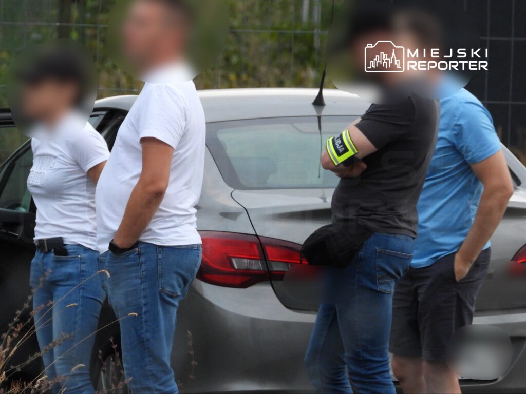 Grupa osób w odzieży roboczej stoi obok szarego samochodu, obserwując otoczenie.