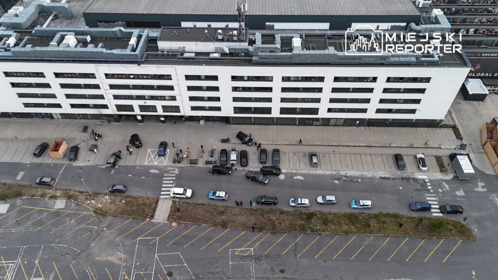 Grupa ludzi z samochodami na parkingu przed nowoczesnym biurowcem, w tle policyjny radiowóz.