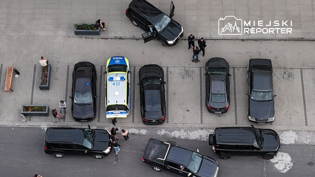 Grupa policjantów rozmawia z mężczyzną na parkingu, obok zaparkowanego radiowozu.