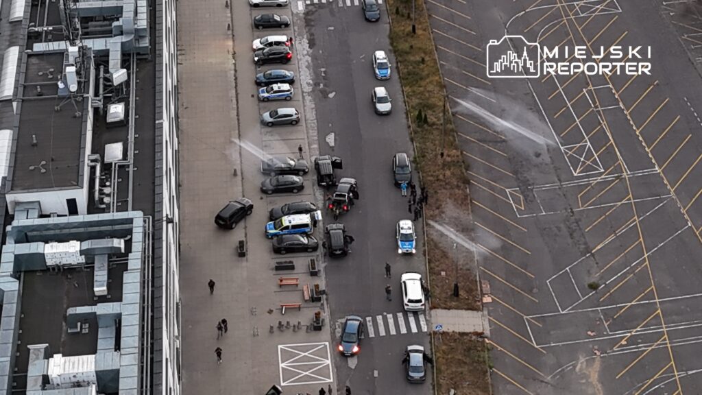 Grupa policjantów kontrolująca pojazdy na ulicy, w tle zaparkowane samochody i nowoczesny budynek.