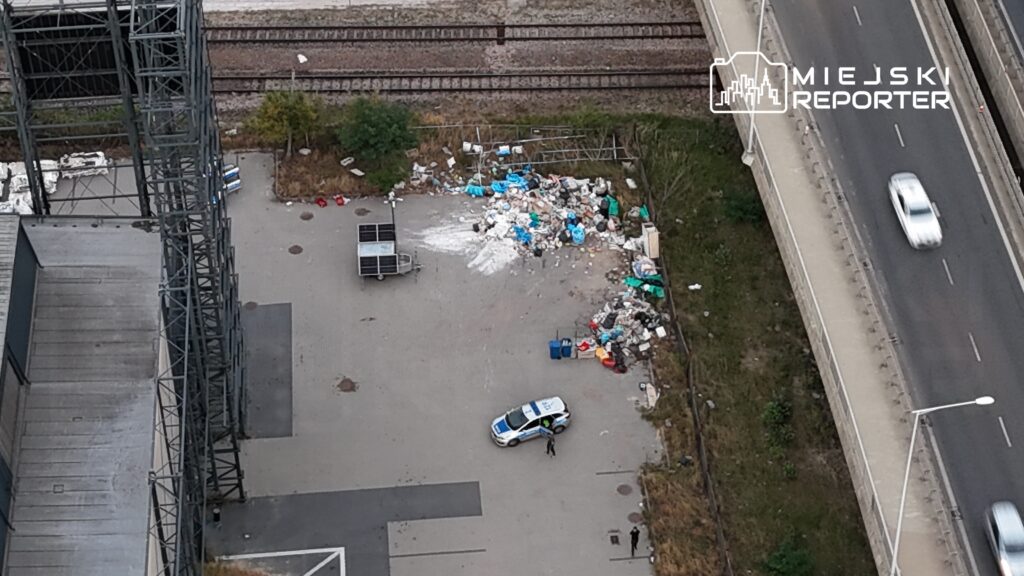 Mundurowy funkcjonariusz przeszukuje teren z porzuconymi odpadami obok drogi i torów kolejowych.