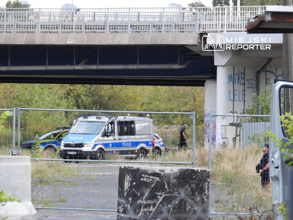 Policyjny radiowóz oraz furgonetka stoją na terenie ogrodzonym, w tle widać graffiti na betonowych ścianach.