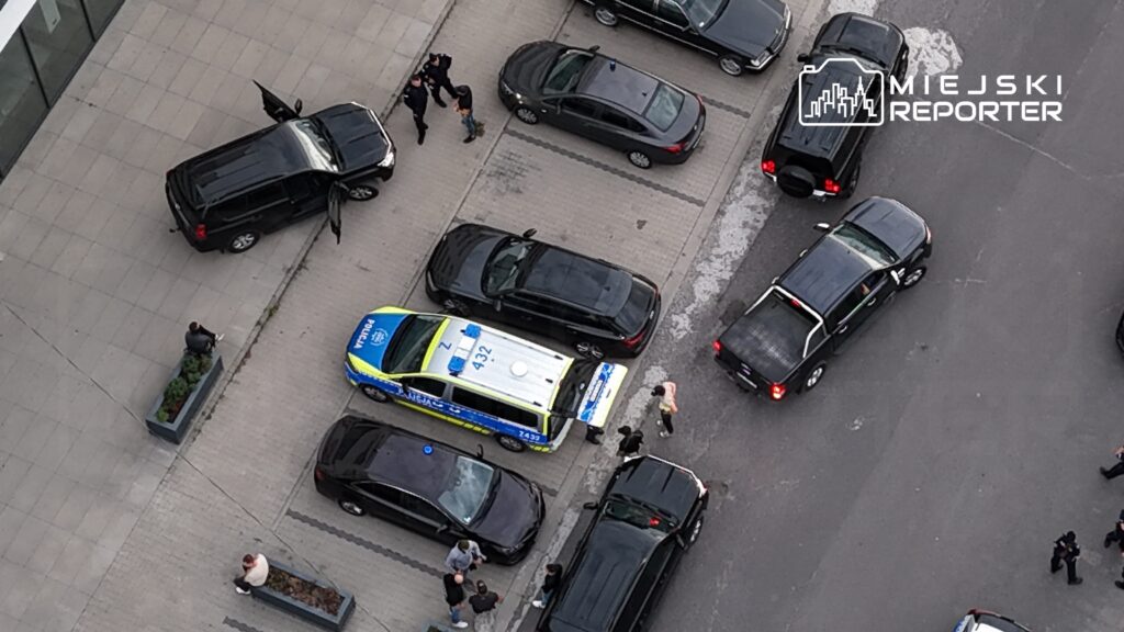 Policyjny radiowóz zatrzymany na parkingu, obok grupka osób rozmawiających, a w tle kilka samochodów osobowych.