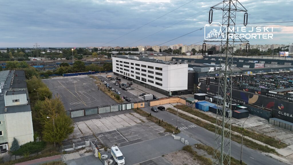 Nowoczesny, biały budynek z dużymi oknami stoi na tle parkingu oraz torów kolejowych w mieście.