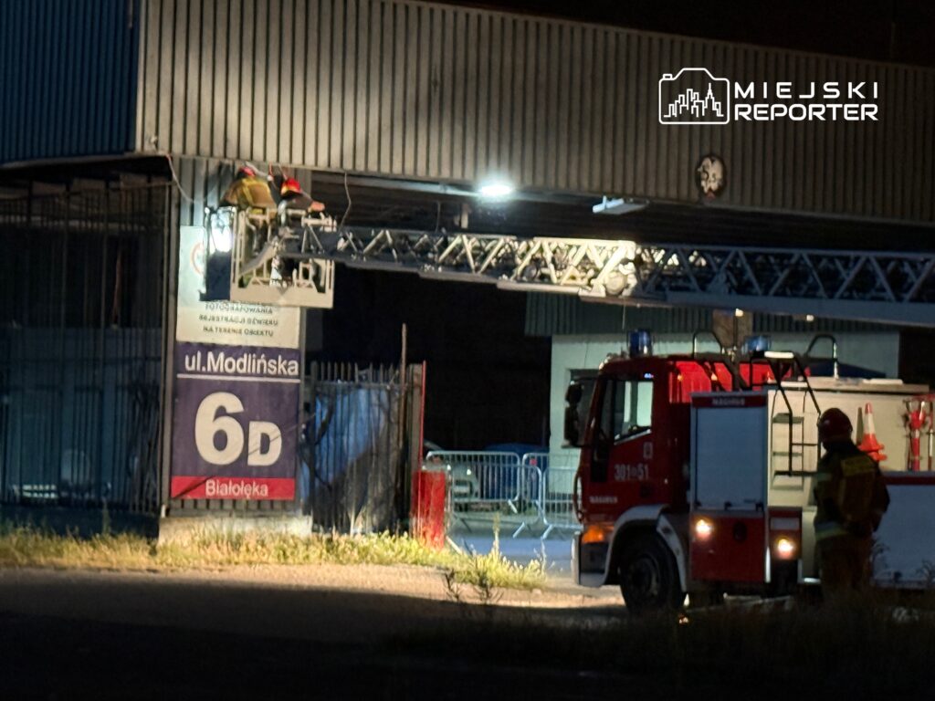 Dwaj strażacy w kaskach pracują na podnośniku, naprawiając oświetlenie nad wejściem do budynku przy ul. Modlińskiej.