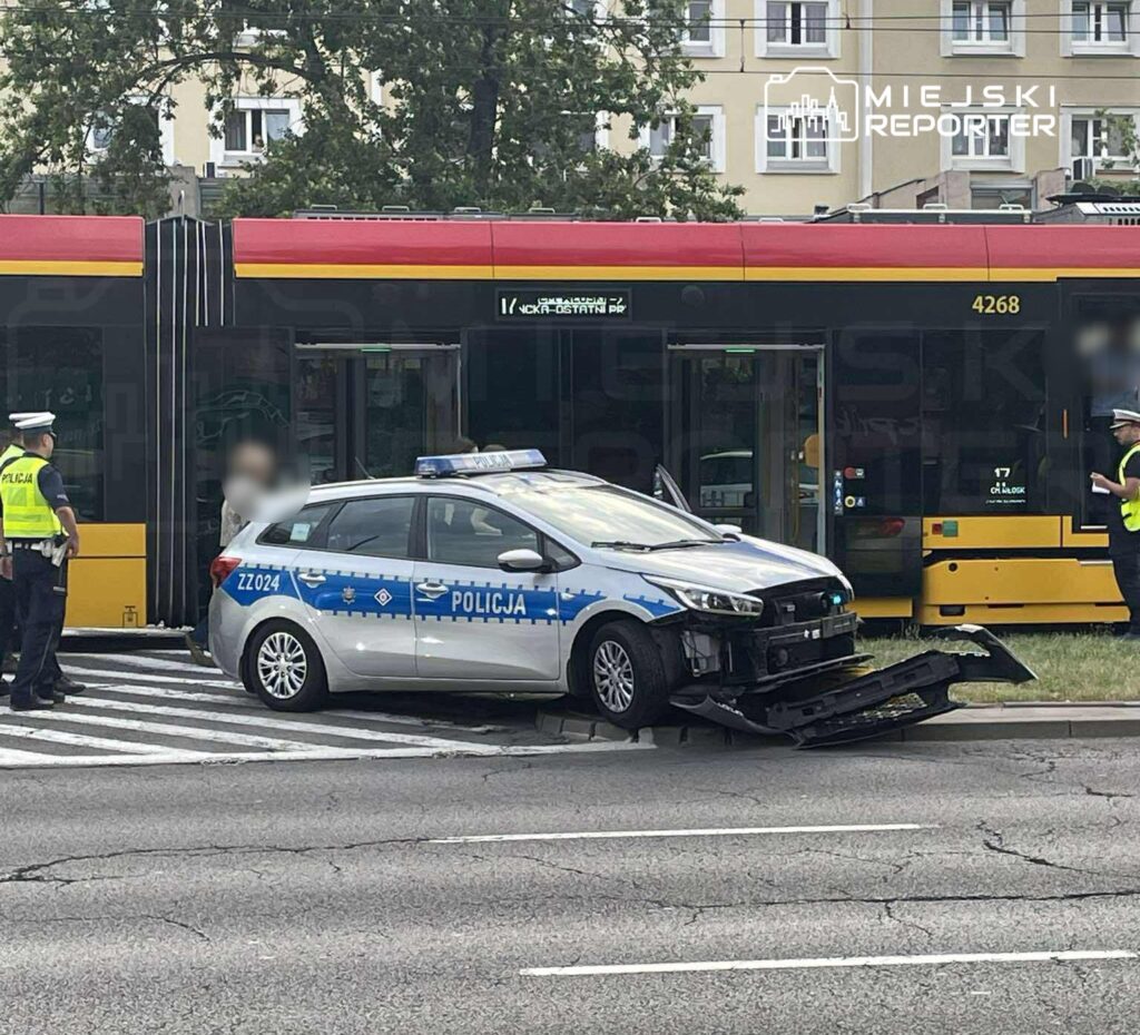 Policyjny radiowóz z uszkodzonym przodem stoi na skrzyżowaniu obok przystanku tramwajowego, z funkcjonariuszami w tle.