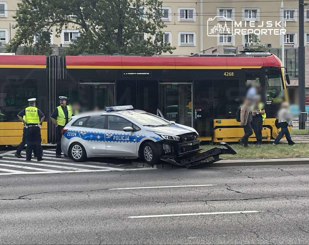 Policyjny radiowóz z uszkodzonym przodem stoi obok przystanku, a funkcjonariusze rozmawiają z pasażerami tramwaju.