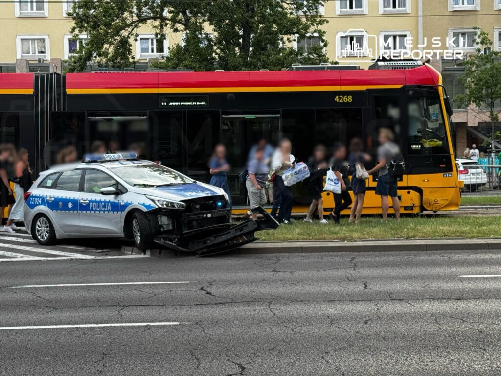 Policyjny radiowóz z uszkodzoną przednią częścią stoi obok tramwaju, a przechodnie czekają na przystanku.