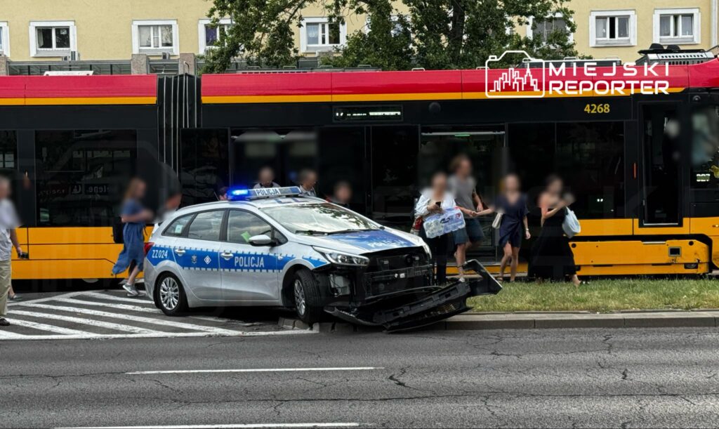Policyjny radiowóz z uszkodzoną przednią częścią stoi na pasie drogowym obok tramwaju, z pasażerami wysiadającymi na przystanku.