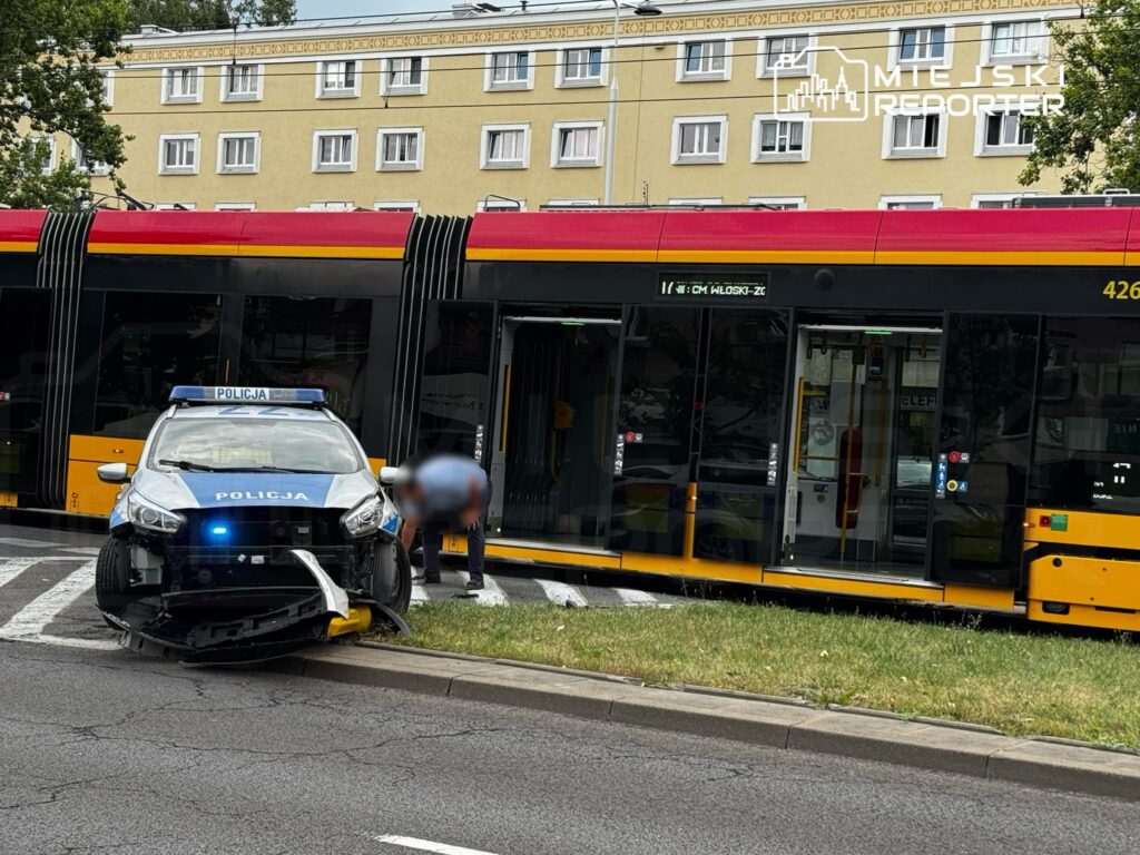 Policyjny samochód z uszkodzoną przednią częścią stoi obok przystanku tramwajowego, gdzie pasażerowie czekają na tramwaj.