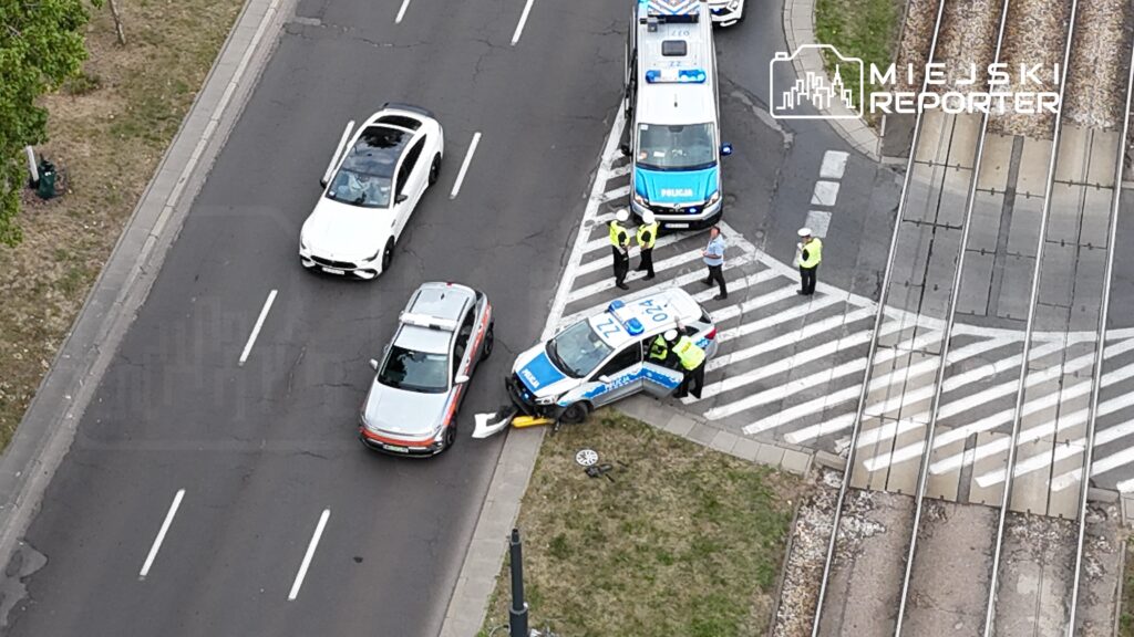 Grupa policjantów rozmawia z kierowcą na skrzyżowaniu, obok stoi policyjny radiowóz z włączonymi sygnałami.