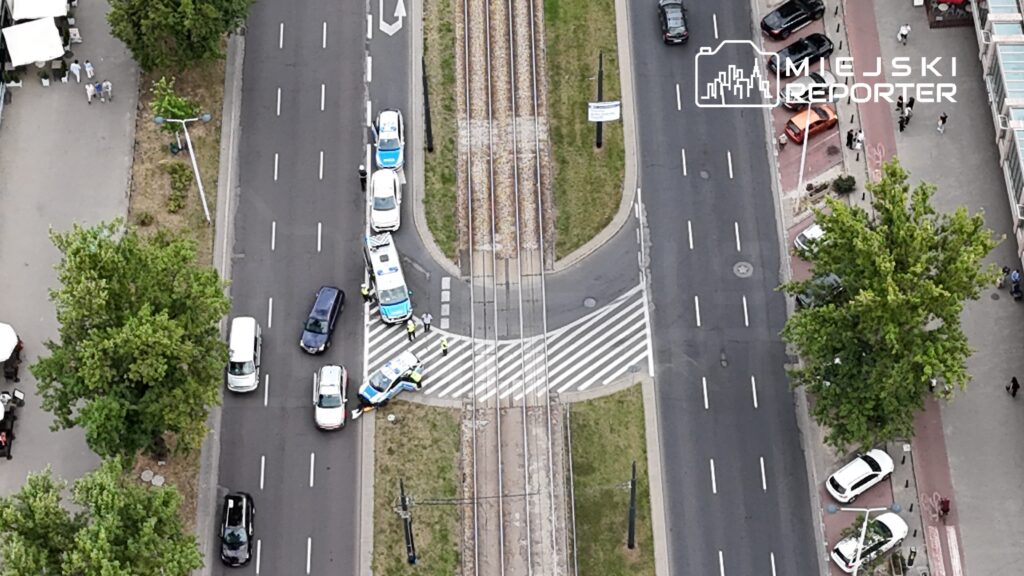 Grupa policjantów kieruje ruchem drogowym na skrzyżowaniu z torami tramwajowymi, w pobliżu zatłoczonej ulicy.