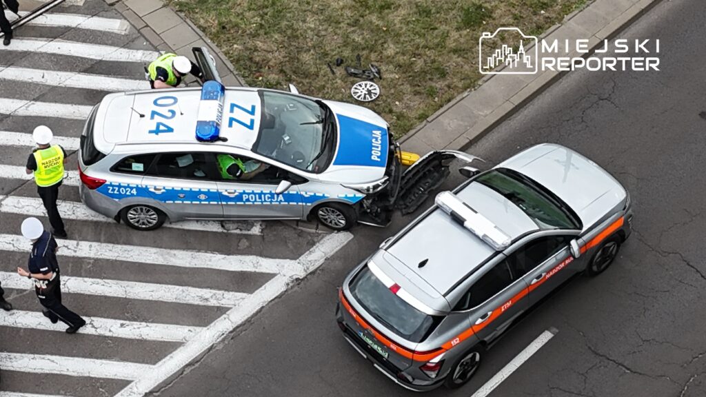 Policjanci w mundurach prowadzą czynności na przejściu dla pieszych obok uszkodzonego radiowozu.