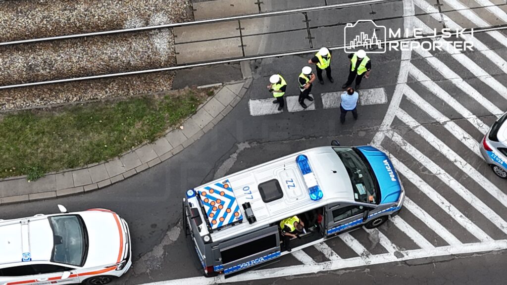 Grupa policjantów w odblaskowych kamizelkach rozmawia z mężczyzną na przejściu dla pieszych obok radiowozu.