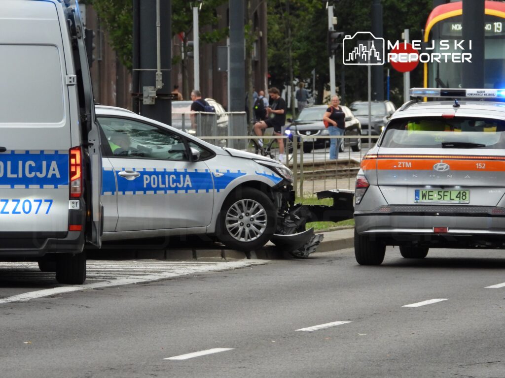 Policyjny samochód z uszkodzoną przednią częścią stoi na chodniku, obok radiowóz z włączonymi sygnałami świetlnymi.