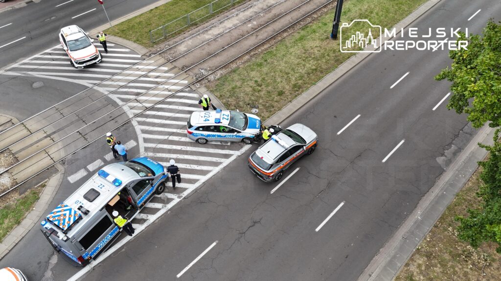 Grupa policjantów rozmawia z kierowcą na skrzyżowaniu, obok zaparkowane pojazdy służbowe.