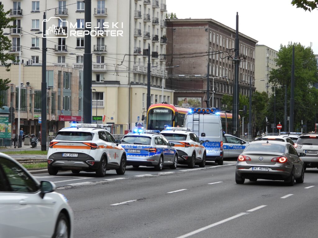 Na ulicy zatrzymane pojazdy, w tym radiowozy z włączonymi sygnałami świetlnymi, obok przejeżdżający autobus.