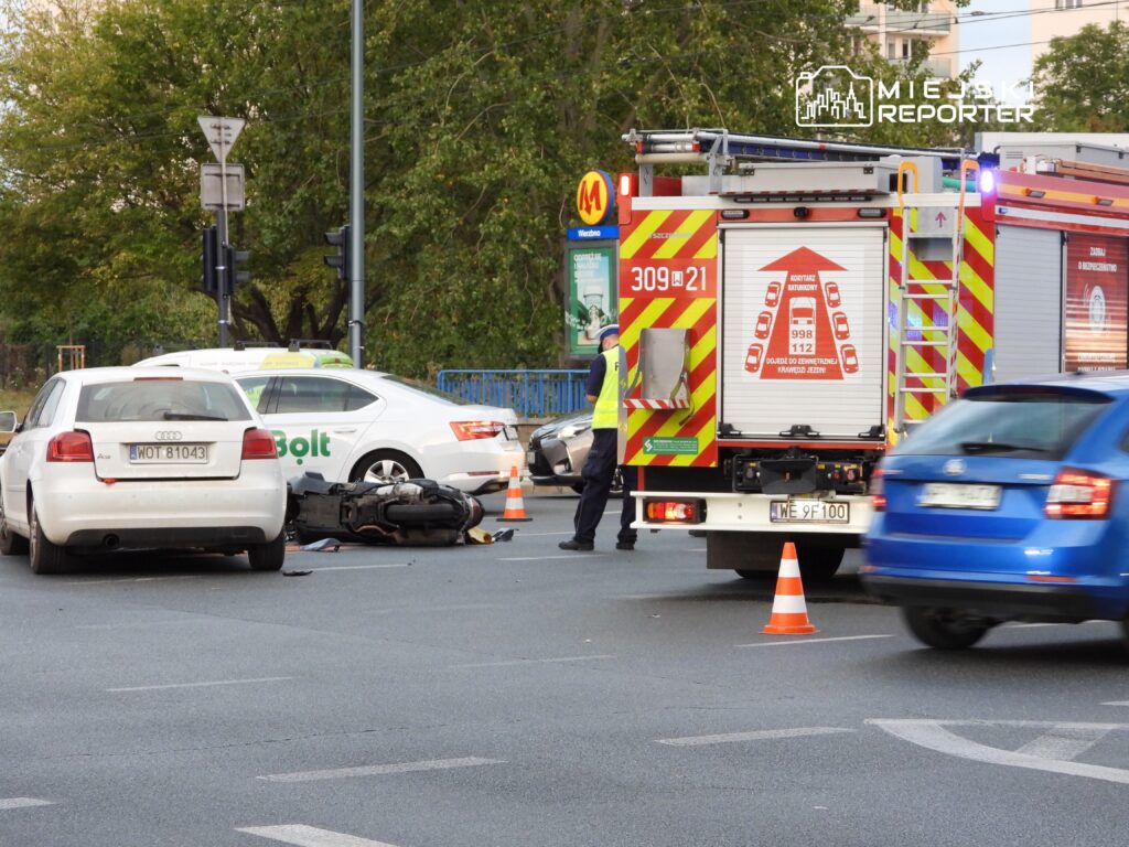 Motocykl leży na jezdni obok dwóch samochodów, a strażak kieruje ruchem w pobliżu wozu strażackiego.
