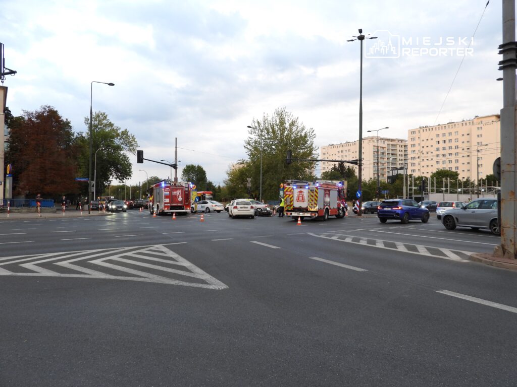Na skrzyżowaniu zatrzymane pojazdy, w tle dwa wozu strażackie z włączonymi sygnałami świetlnymi.