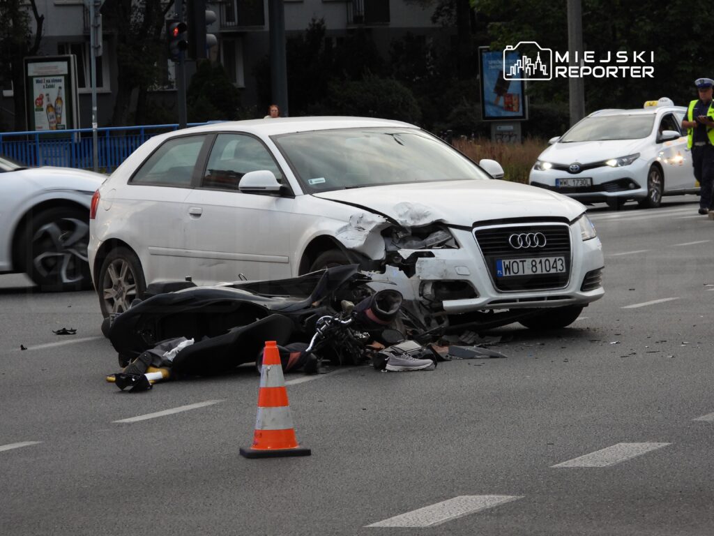 Zderzenie samochodu osobowego z motocyklem na ruchliwej ulicy, wokół widoczne zniszczenia i sygnalizacja świetlna.