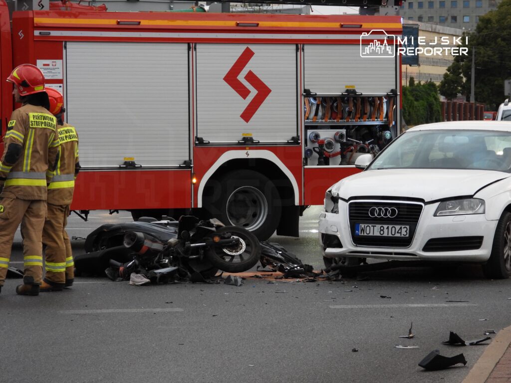 Strażacy w mundurach przy uszkodzonym samochodzie osobowym i rozbitej motocyklu na drodze.