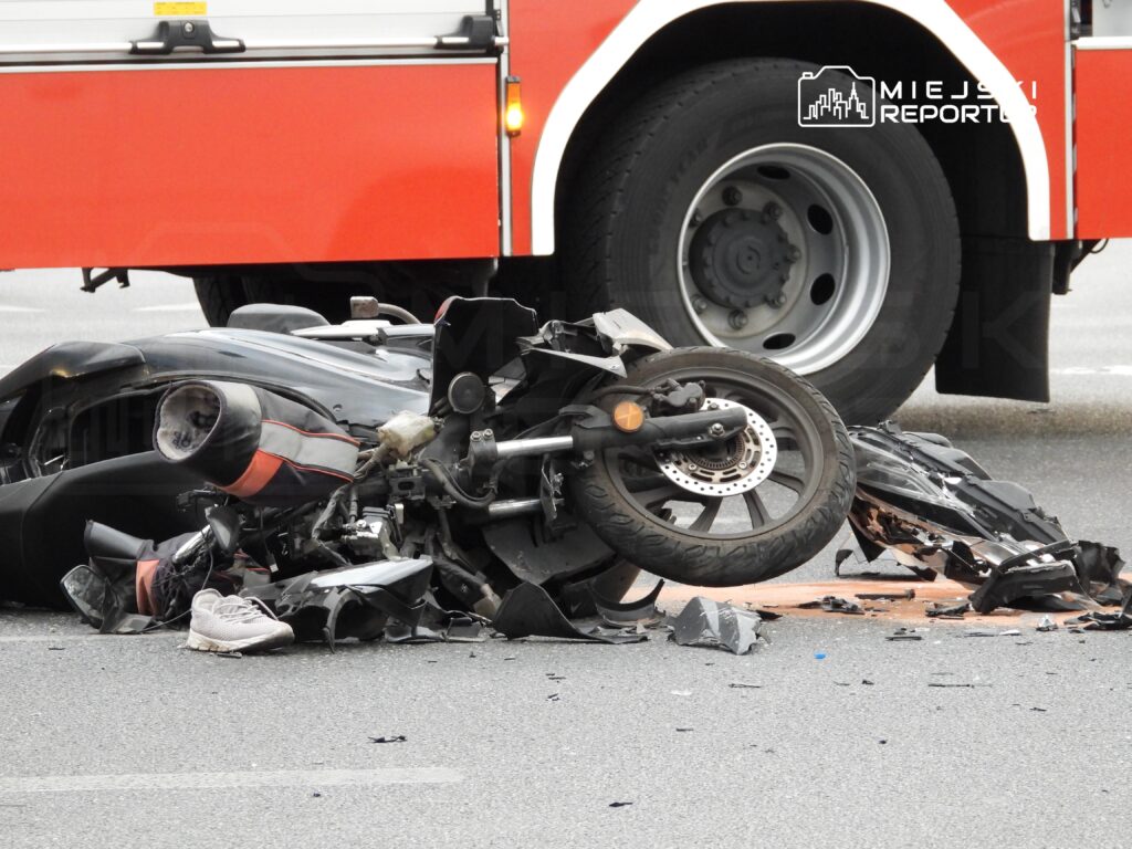 Uszkodzony motocykl leży na jezdni obok czerwonego wozu strażackiego, w tle fragmenty rozbitej karoserii.