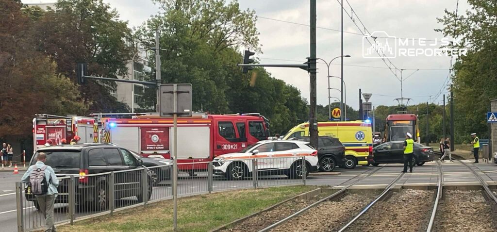 Na skrzyżowaniu ulicy stoją pojazdy ratunkowe, w tym straż pożarna i karetka, otoczone przez policję i gapiów.