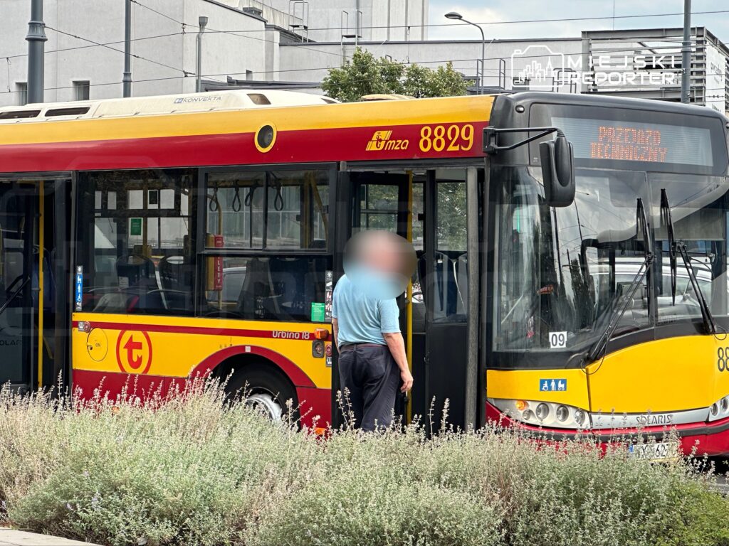 Mężczyzna w niebieskiej koszuli wchodzi do czerwono-żółtego autobusu miejskiego na przystanku.