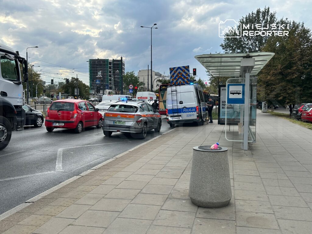 Ruch uliczny z zablokowanymi pojazdami, w tym radiowozem policyjnym, obok przystanku autobusowego.