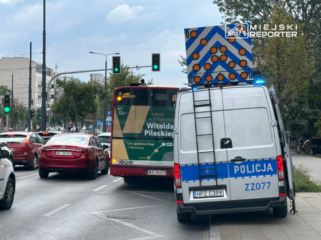 Policyjny van z włączonymi sygnałami świetlnymi zatrzymany przy ruchliwej ulicy, obok autobusu miejskiego.