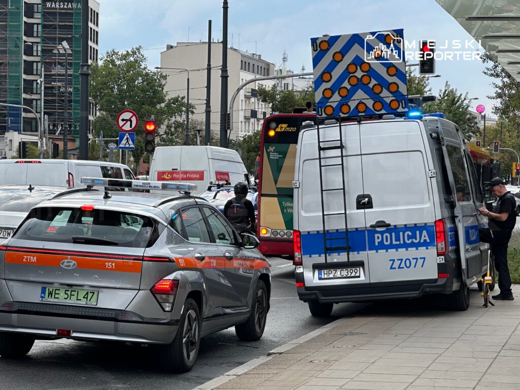 Radiowóz policyjny zatrzymany na ulicy, obok samochodu elektrycznego ZTM, w tle czerwone światło i ruch uliczny.