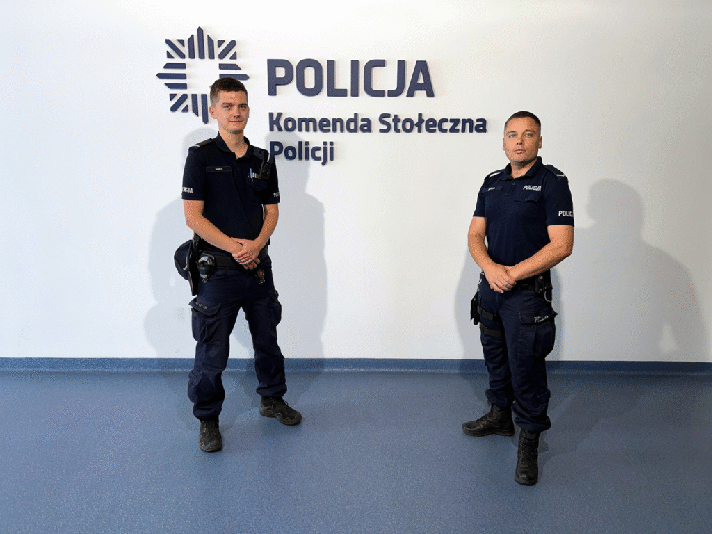 Dwaj policjanci w mundurach stoją obok siebie w nowoczesnym wnętrzu komendy policji.
