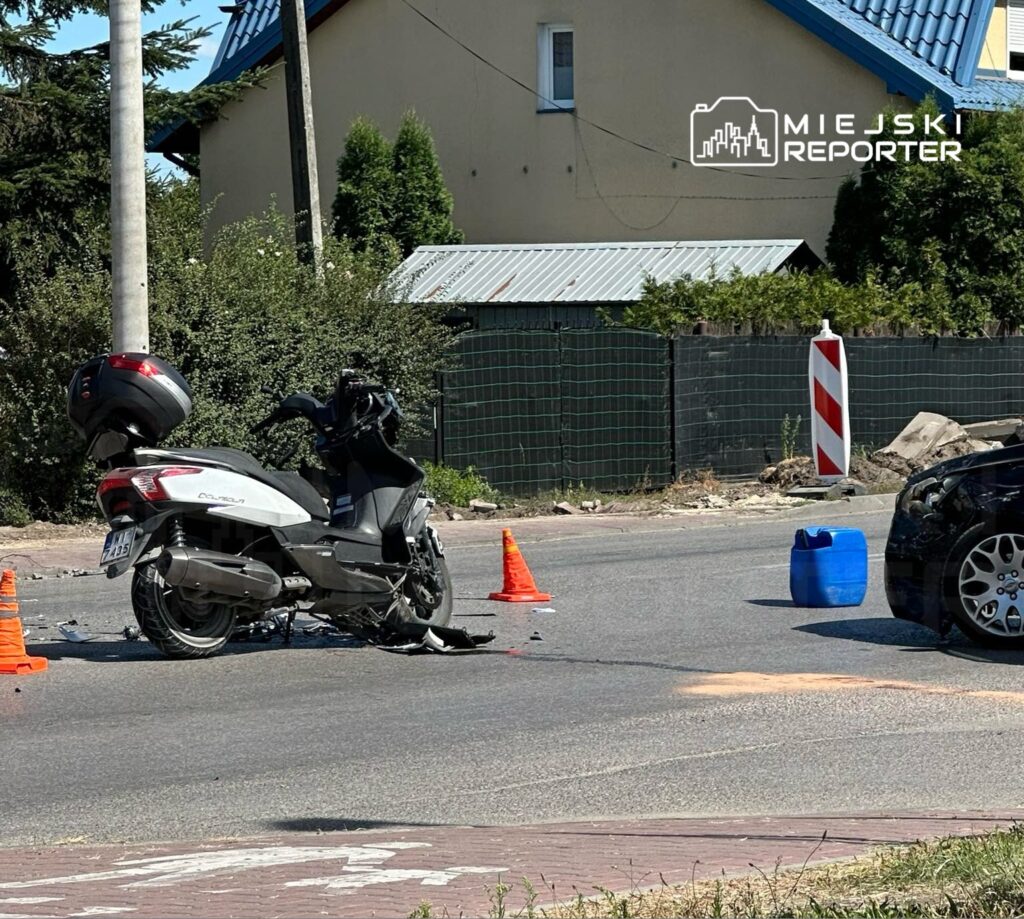 Skuter leży na boku obok drogi, otoczony pomarańczowymi pachołkami, a w tle widać czarny samochód i budynek mieszkalny.