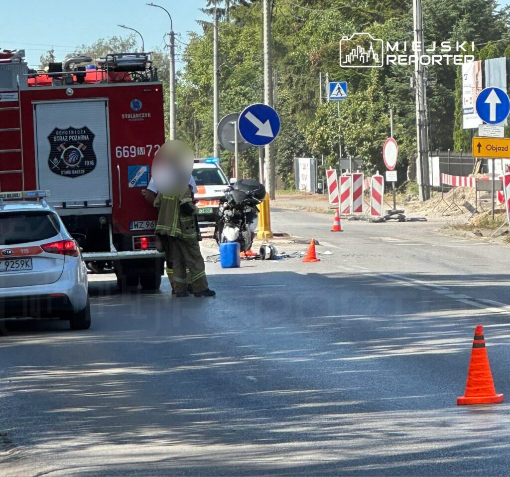 Strażak w mundurze stoi obok wozu strażackiego na drodze, obok leży motocykl i trwają prace zabezpieczające miejsce zdarzenia.