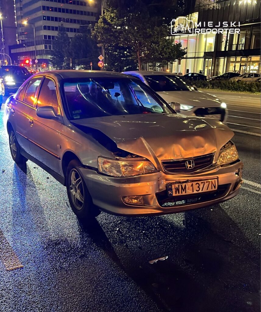 Uszkodzony samochód osobowy marki Honda z wgniecionym przodem stoi na mokrej jezdni w miejskim otoczeniu.