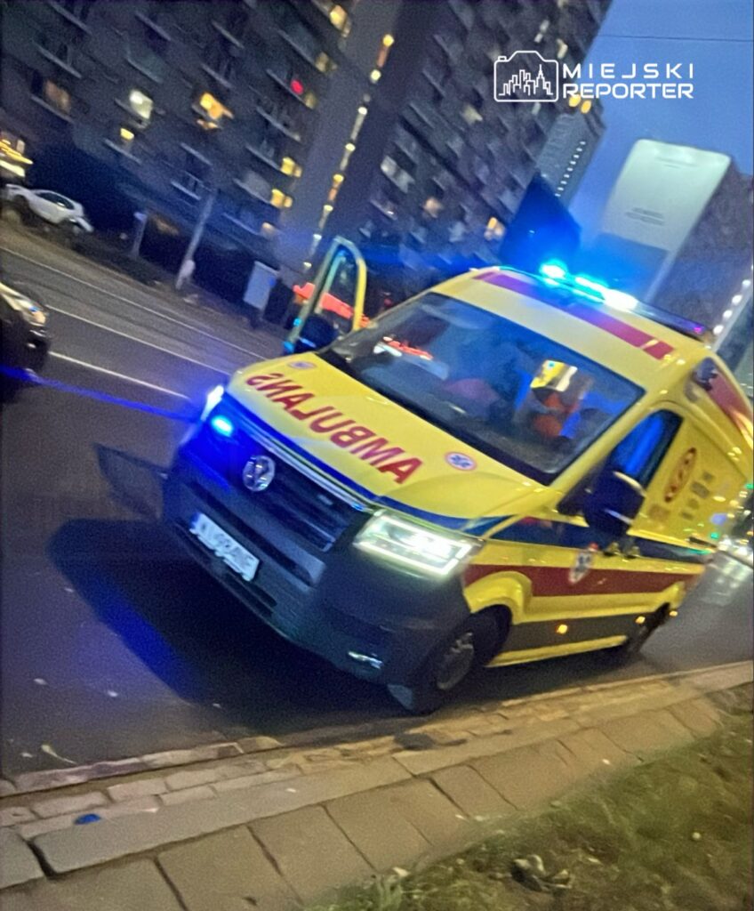 Karetka pogotowia z włączonymi sygnałami świetlnymi zatrzymana na ulicy w pobliżu wysokiego budynku mieszkalnego.
