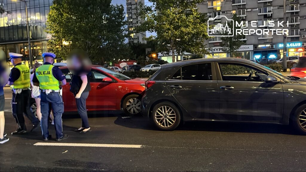 Policjanci rozmawiają z kobietą przy zderzonych samochodach na ulicy w mieście, w tle widoczne oświetlone budynki.