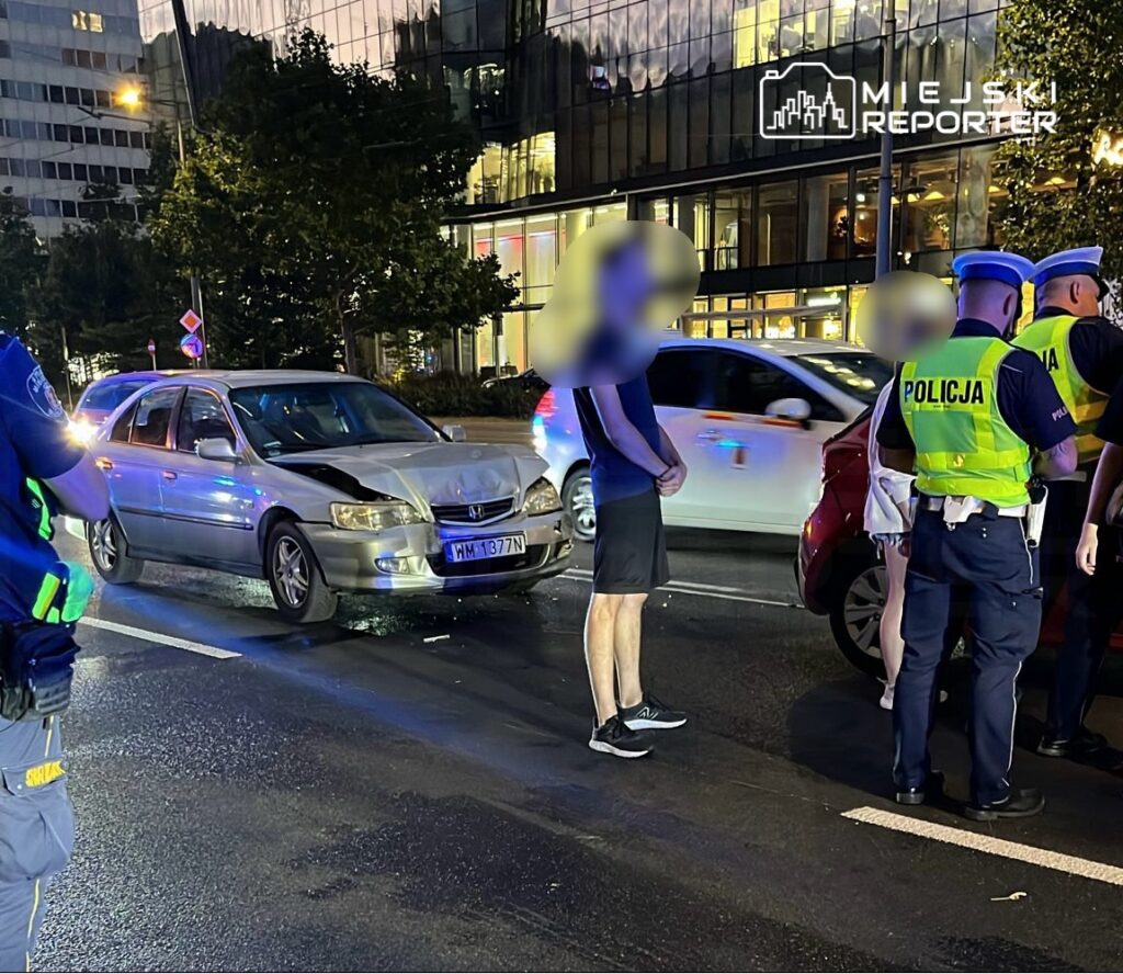 Kierowca z uszkodzonym samochodem rozmawia z policjantami na ruchliwej ulicy w nocy. W tle nowoczesne budynki.