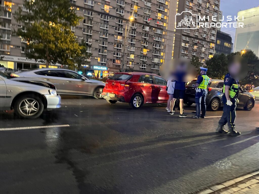 Policjanci rozmawiają z uczestnikami kolizji drogowej na ruchliwej ulicy, obok uszkodzonych pojazdów.