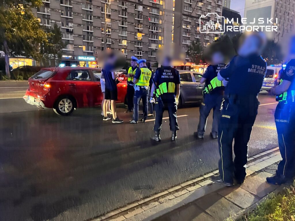 Grupa funkcjonariuszy policji i straży miejskiej rozmawia z kierowcą czerwonego samochodu na ulicy.