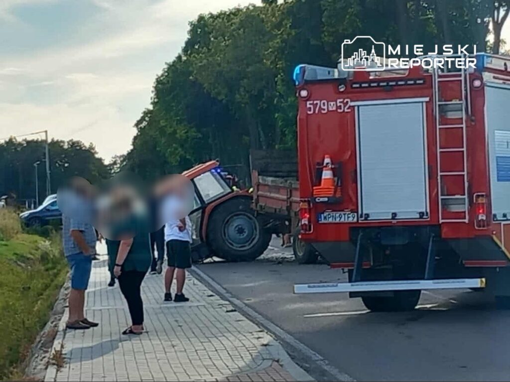 Grupa osób stoi na chodniku obok przewróconego traktora, obok parkowany wóz strażacki.