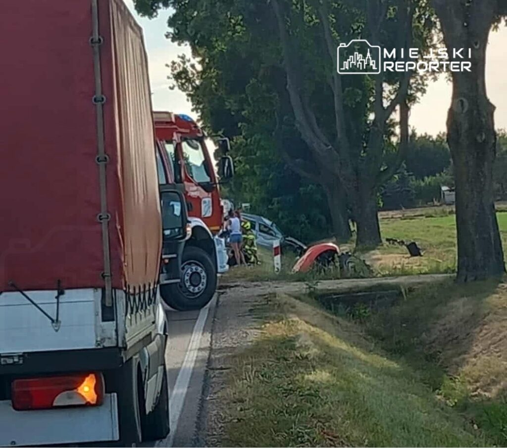 Kierowca w białej koszulce rozmawia z ratownikami przy uszkodzonym samochodzie na poboczu drogi, obok stoi straż pożarna.
