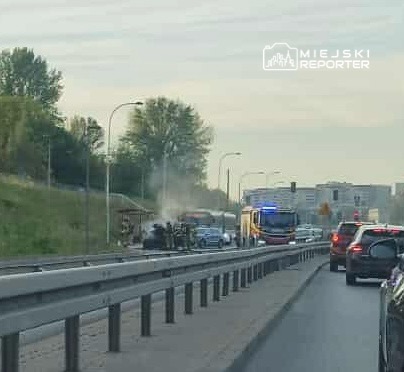 Na drodze widać samochód objęty dymem, obok zatrzymane pojazdy oraz straż pożarną z włączonymi sygnałami świetlnymi.