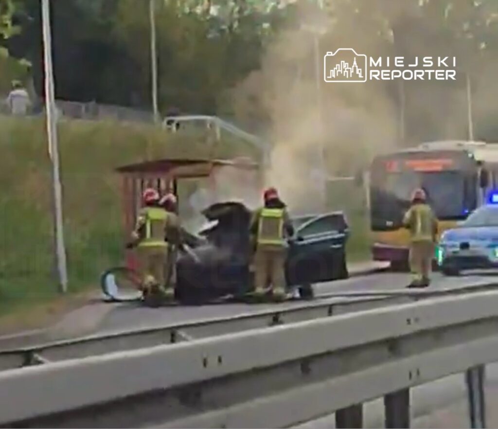 Strażacy gaszą pożar samochodu na poboczu drogi, obok przystanku i przejeżdżającego autobusu.