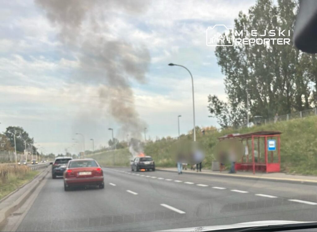 Samochód na poboczu drogi płonie, wydobywający się z niego dym unosi się w powietrze; w tle widać przystanek autobusowy.