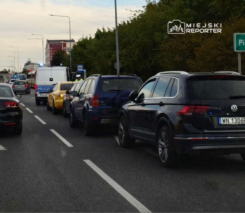 Kolejka samochodów zatrzymanych na drodze, z radiowozem policji w tle oraz sygnalizacją świetlną.