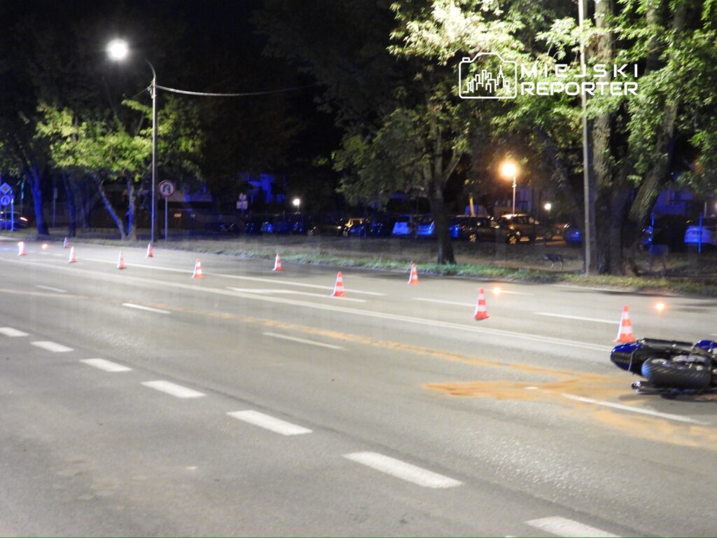 Na jezdni leży przewrócony motocykl, a wokół niego rozstawione są pachołki ostrzegawcze. W tle widać oświetlone drzewa i samochody.