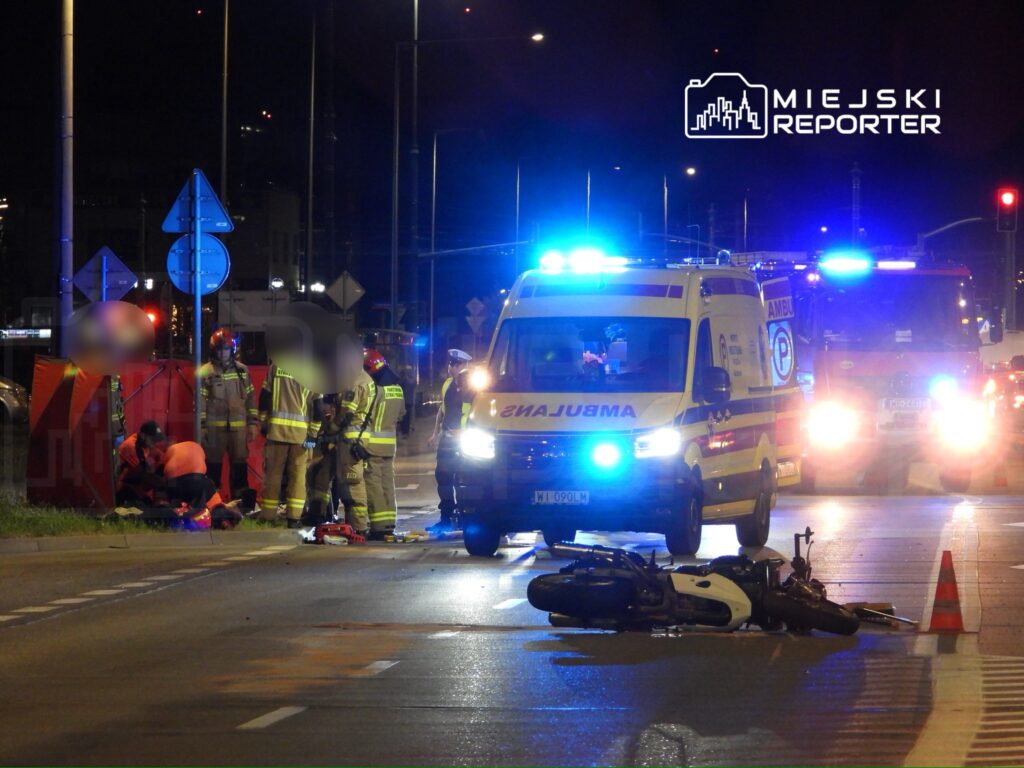 Grupa ratowników medycznych udziela pomocy poszkodowanemu na jezdni obok przewróconego motocykla w nocy.