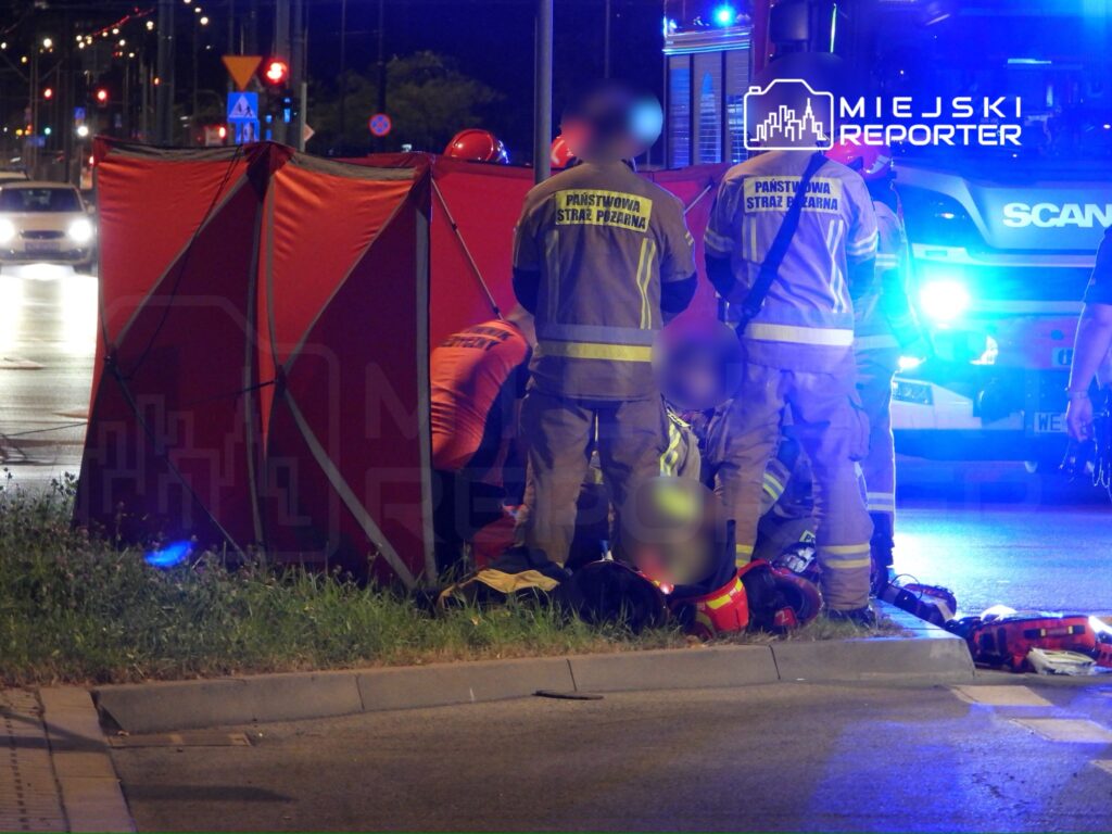Grupa strażaków w mundurach, pracująca przy wypadku drogowym, otoczona czerwonym namiotem, w nocy na ulicy.